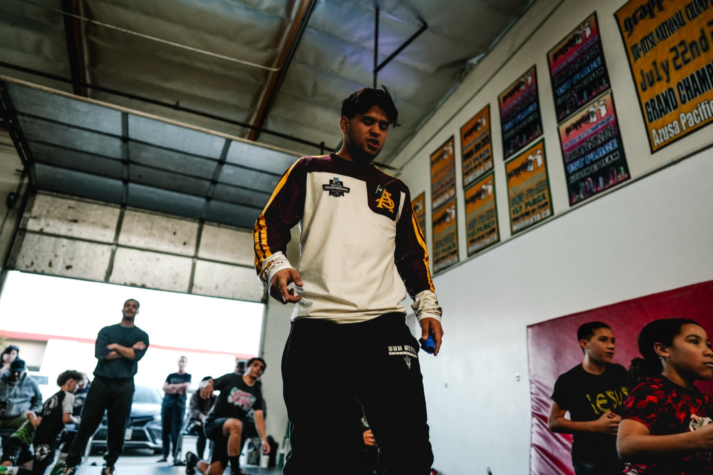A coach in a maroon and white Arizona State warmup top and black sweatpants walks across a gym floor talking, with tournament posters on the wall behind him and young wrestlers training in the background.