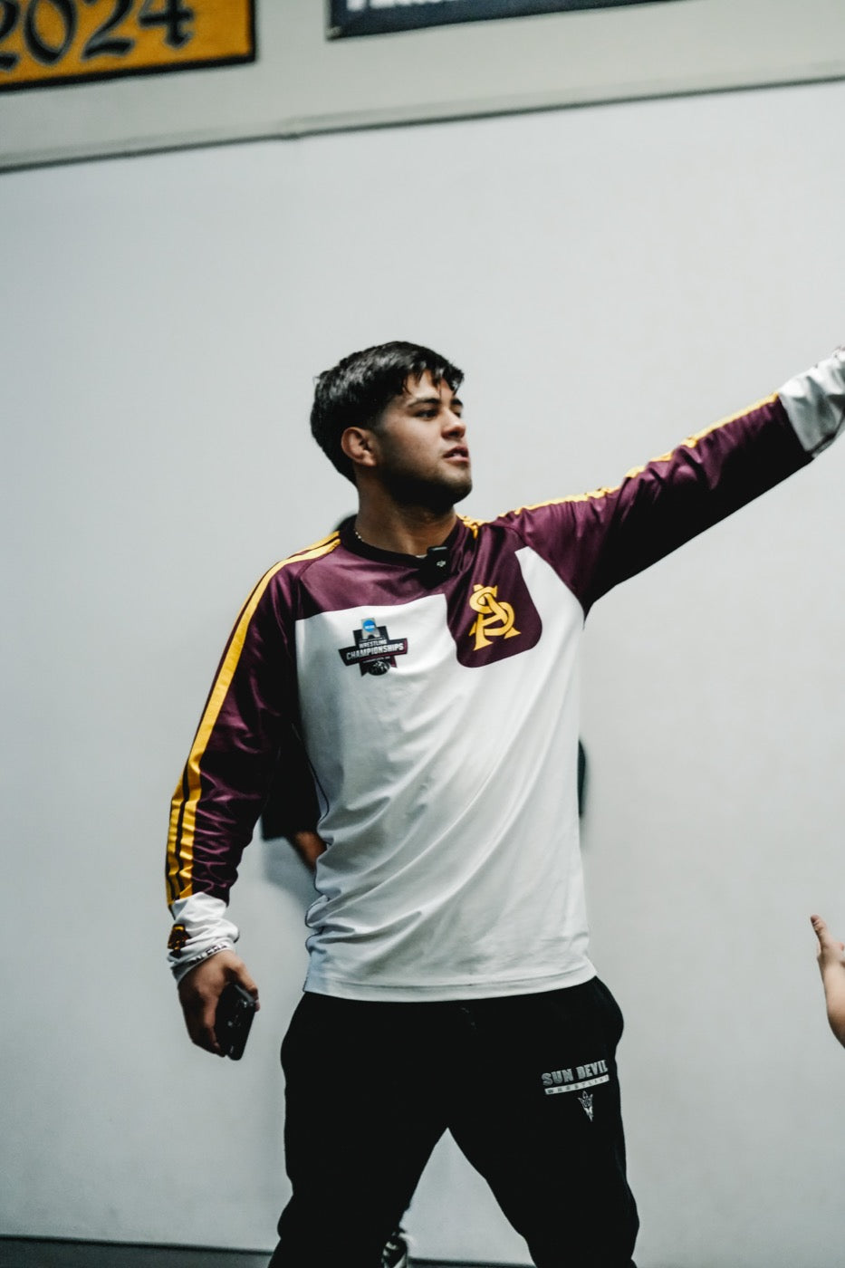 Jesse Vasquez in a maroon, white and gold Arizona State Sun Devils warm-up top with the interlocking AS logo and an NCAA Wrestling Championships patch, arm extended to his left, holding a phone, standing against a plain wall.